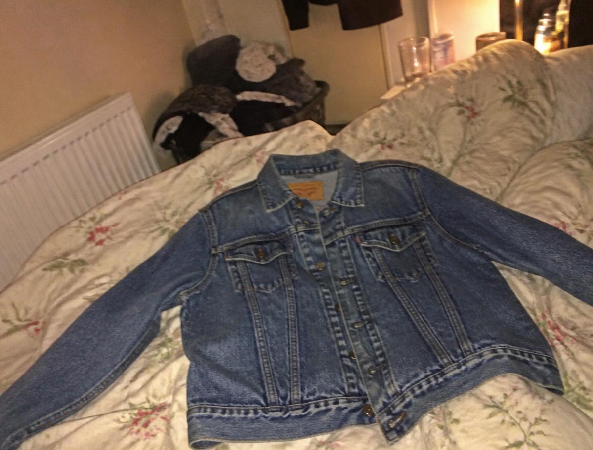 A denim jacket photographed on a messy bed in dim bedroom light