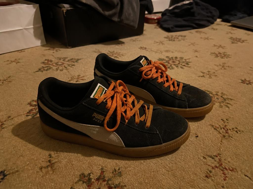 Black suede trainers with orange laces on a patterned bedroom carpet in dim light